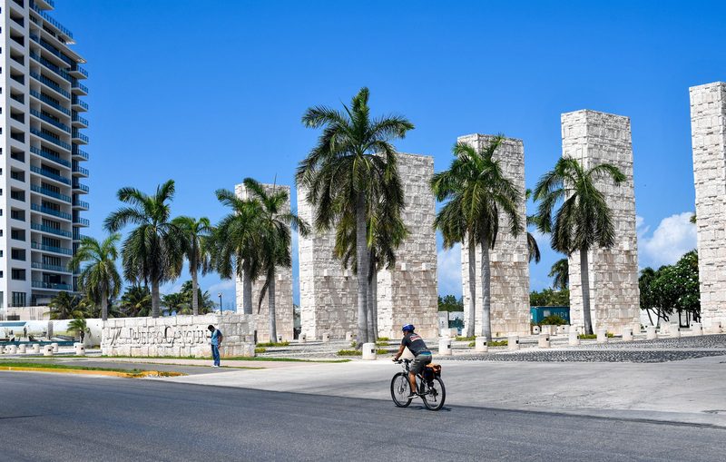 La entrada de Puerto Cancún: columnas de piedra, el muro rotulado, palmeras y una torre detrás.
